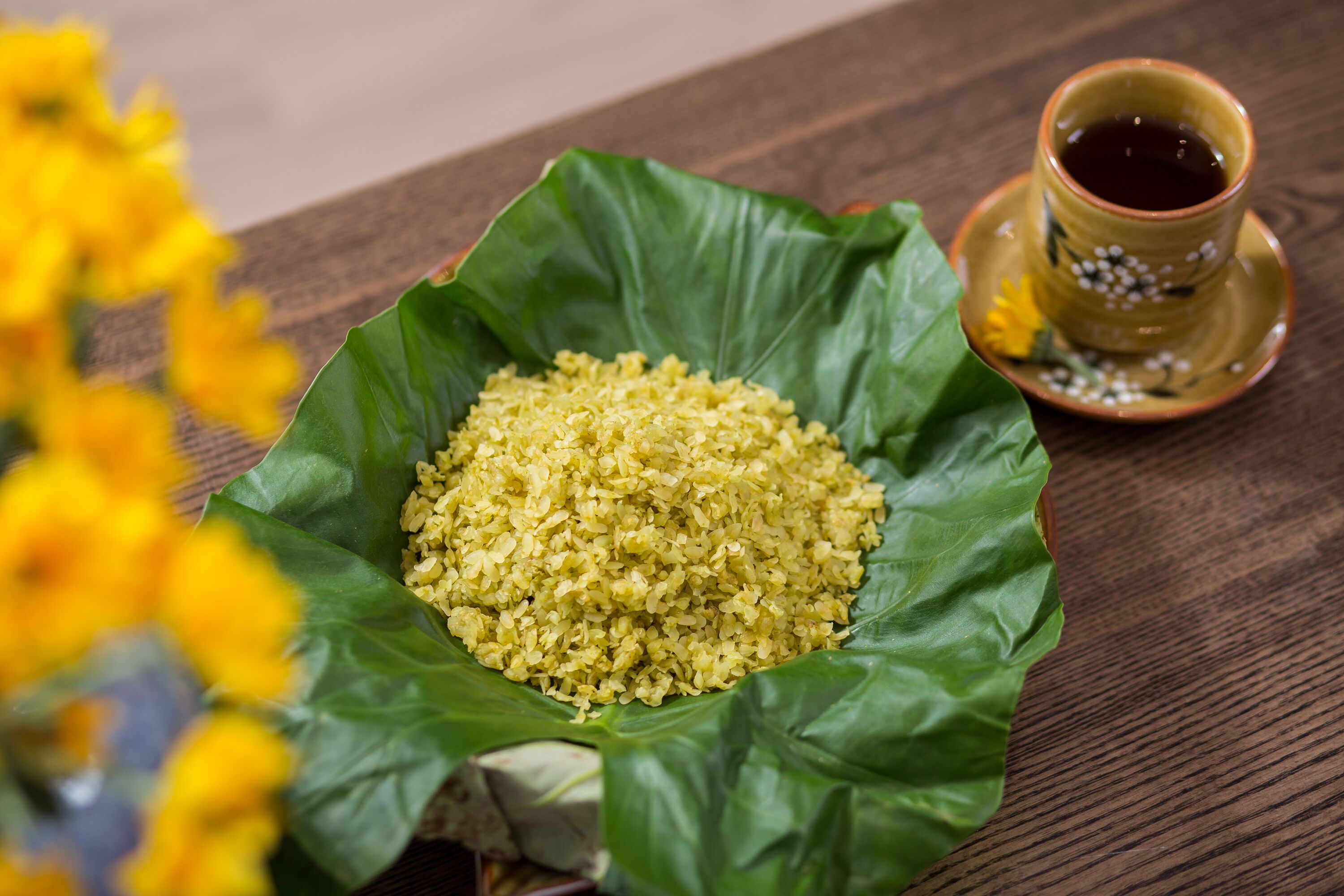 Un bol de riz jaune posé sur une feuille verte, une tasse de thé et une table en bois.