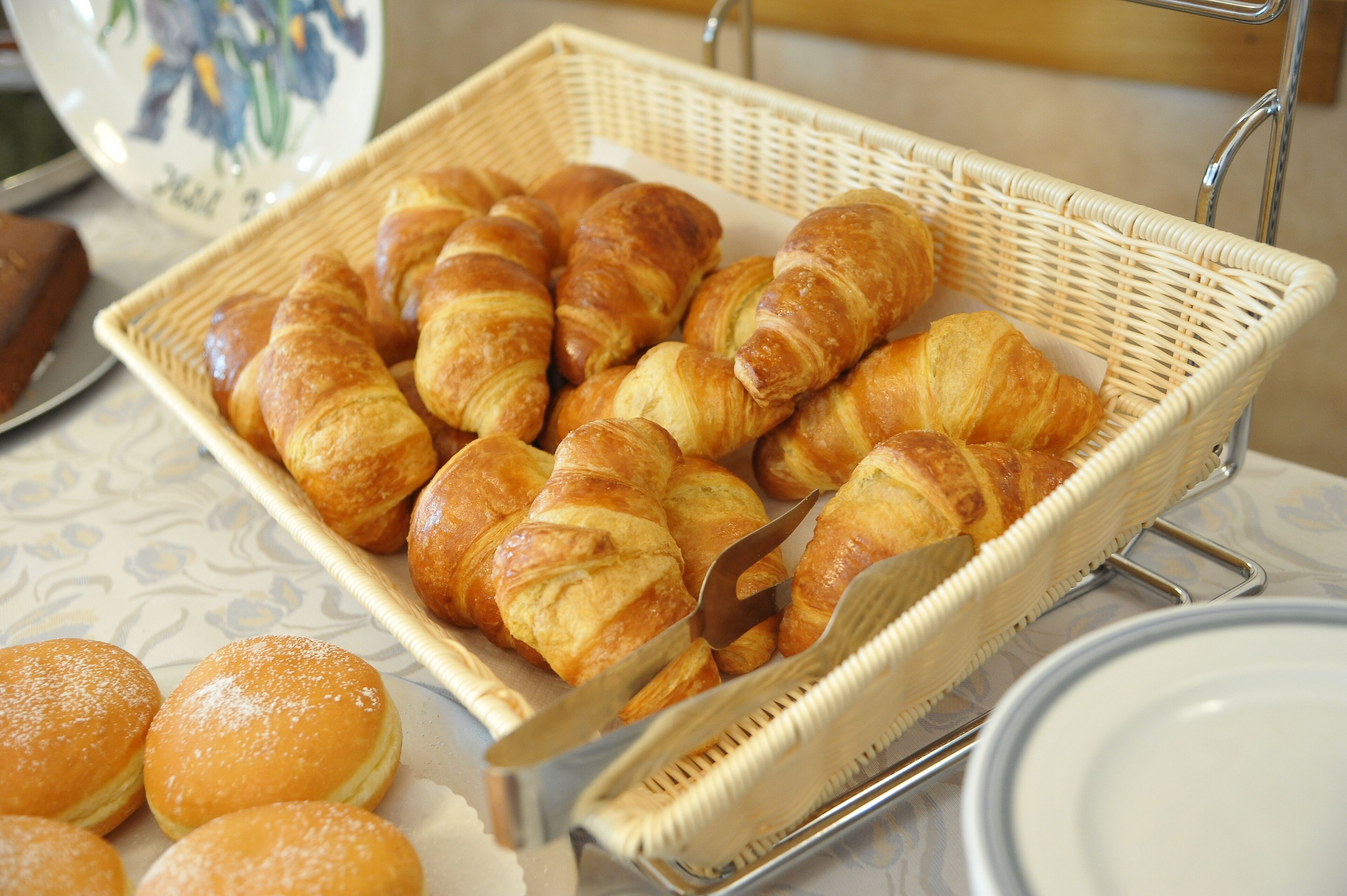 A wicker basket filled with golden-brown croissants and a few round pastries, placed on a table with a patterned tablecloth.