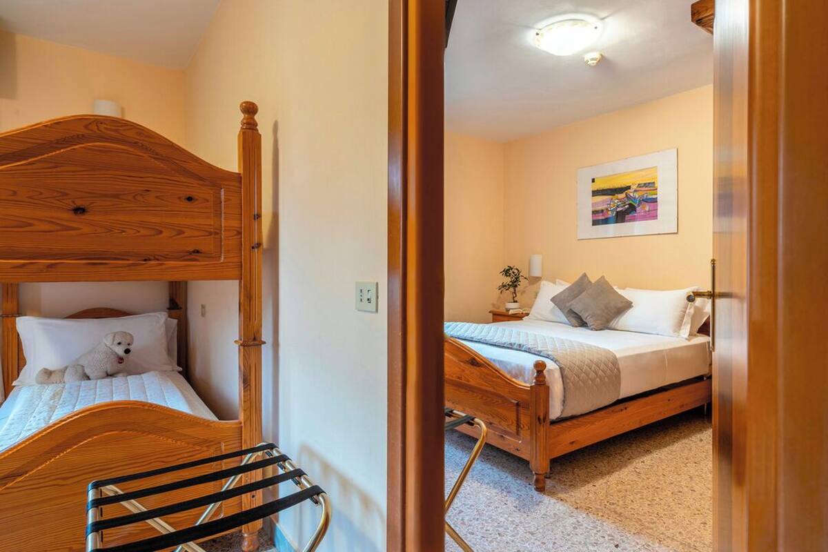 A bedroom with a bunk bed and a single bed, both with wooden frames. There is a small table with a plant and a lamp on the single bed.