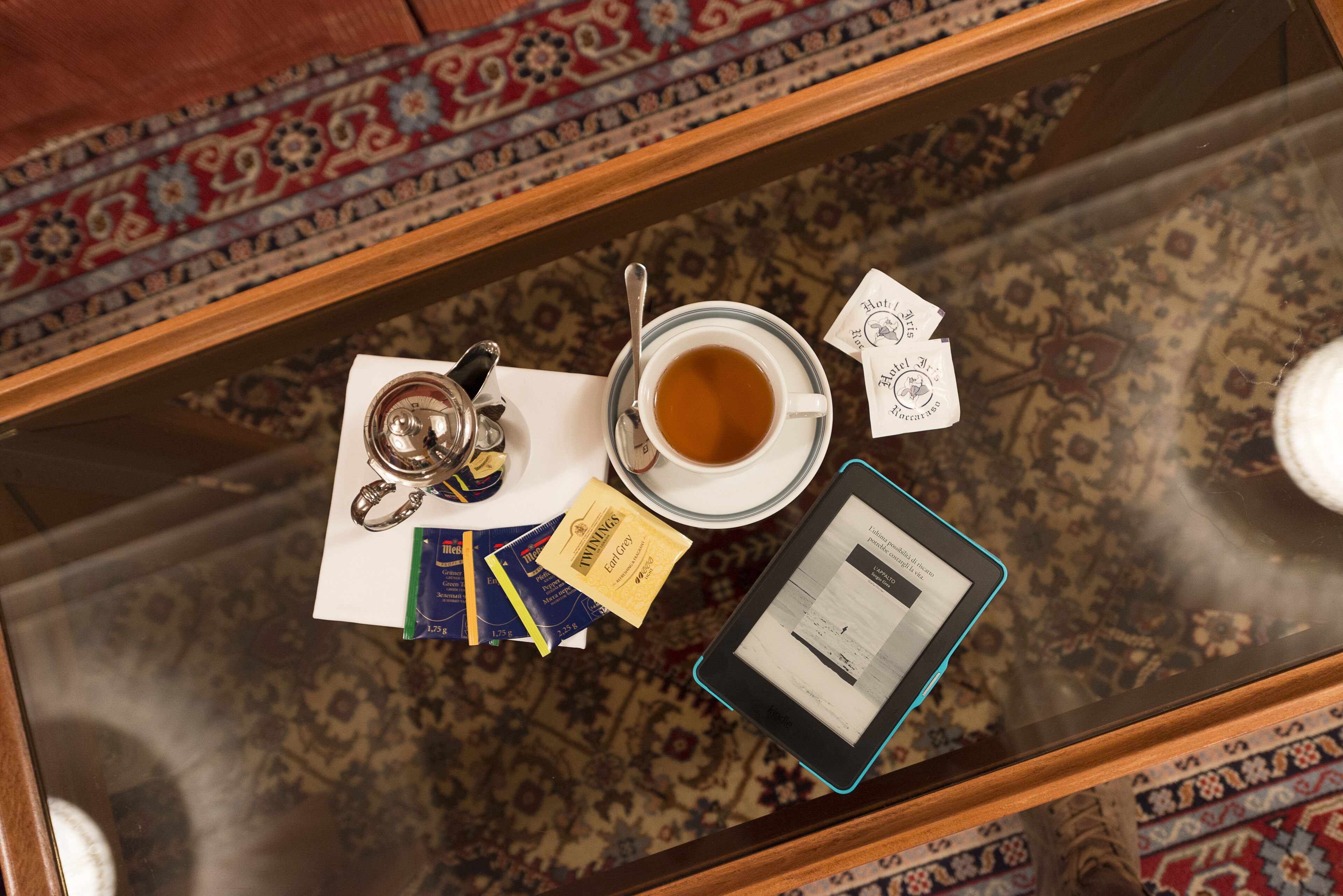 Un tavolino di vetro con una tazza di tè, un tablet, un paio di chiavi, una carta e un piccolo pacchetto su un tappeto decorato.