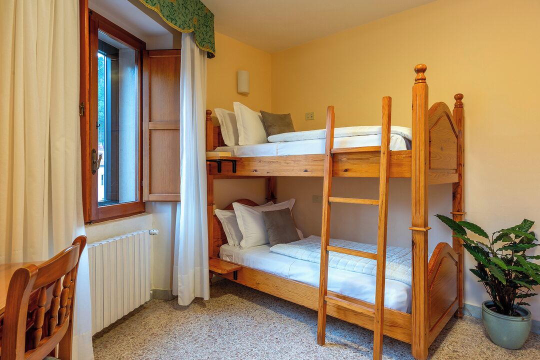 Camera con letto a castello, una testiera in legno, una finestra con tende, un termosifone, una sedia e una pianta in vaso.