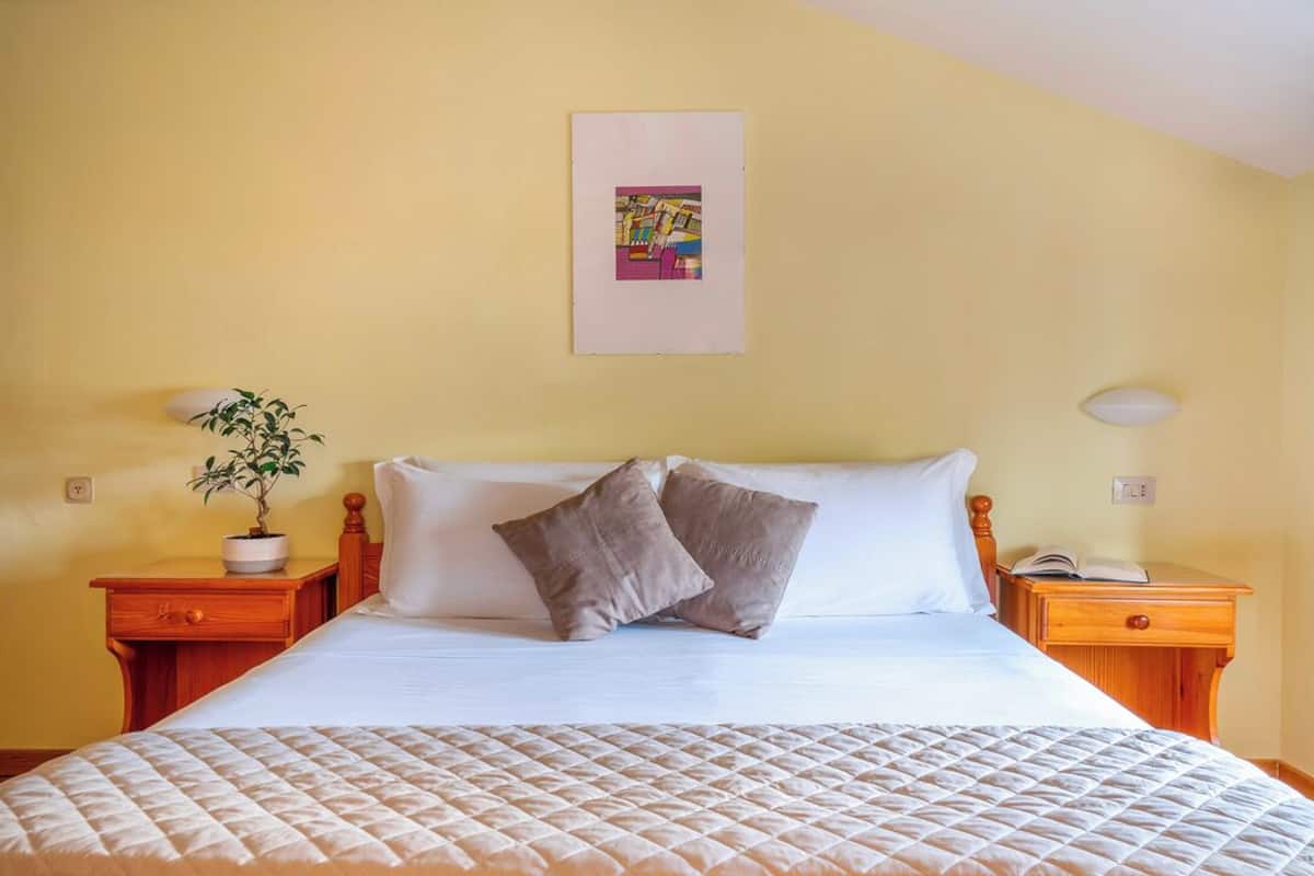 A neatly made bed with a quilted cover, two pillows, a bedside plant, and a framed picture on the wall.