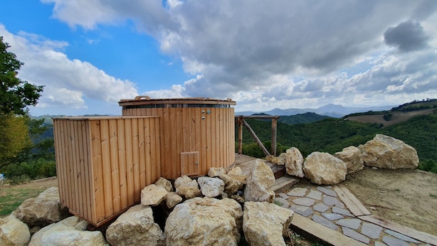 Un letrina de madera con puerta metálica, rodeada de rocas grandes y un sendero de piedra, frente a un paisaje montañoso con cielo despejado.