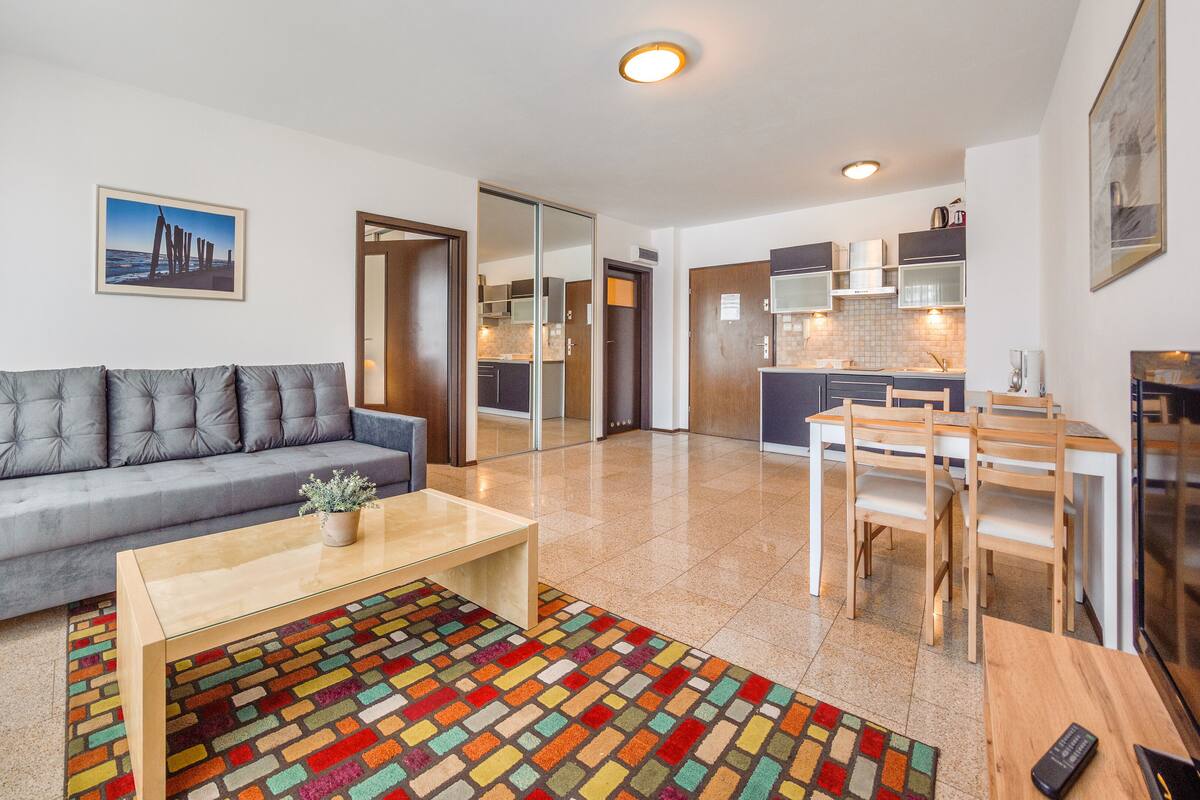 A modern living room with a grey sofa, a wooden coffee table, and a colorful rug. There is a kitchen area with cabinets and a dining table.