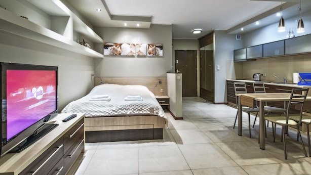Una habitación de hotel moderna con cama, televisor, mesita de noche y una pequeña kitchenette.