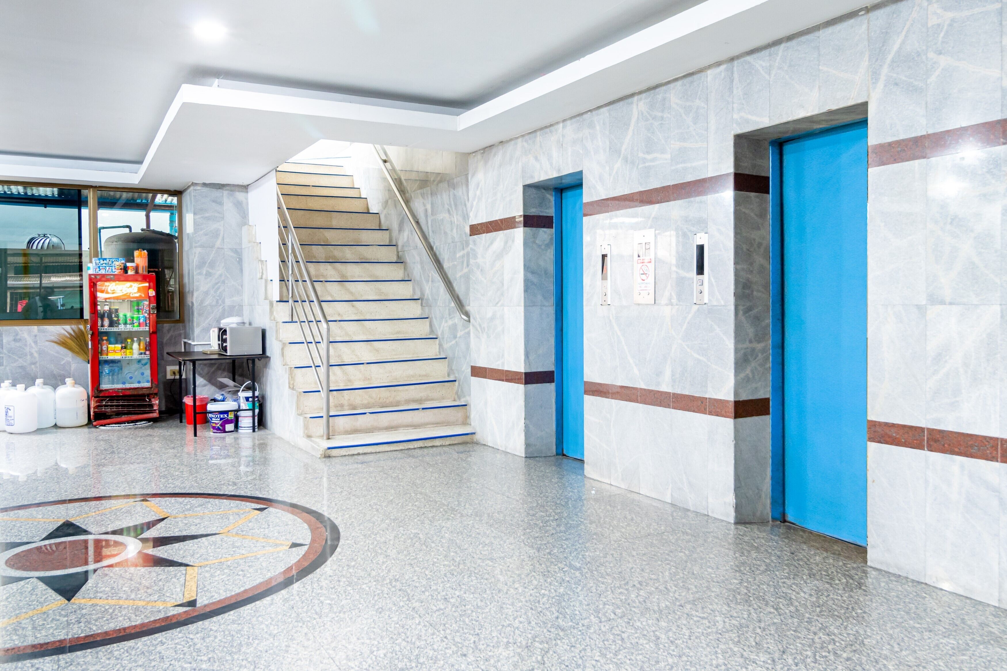 A modern lobby with a marble floor, a vending machine, a staircase, and blue doors.