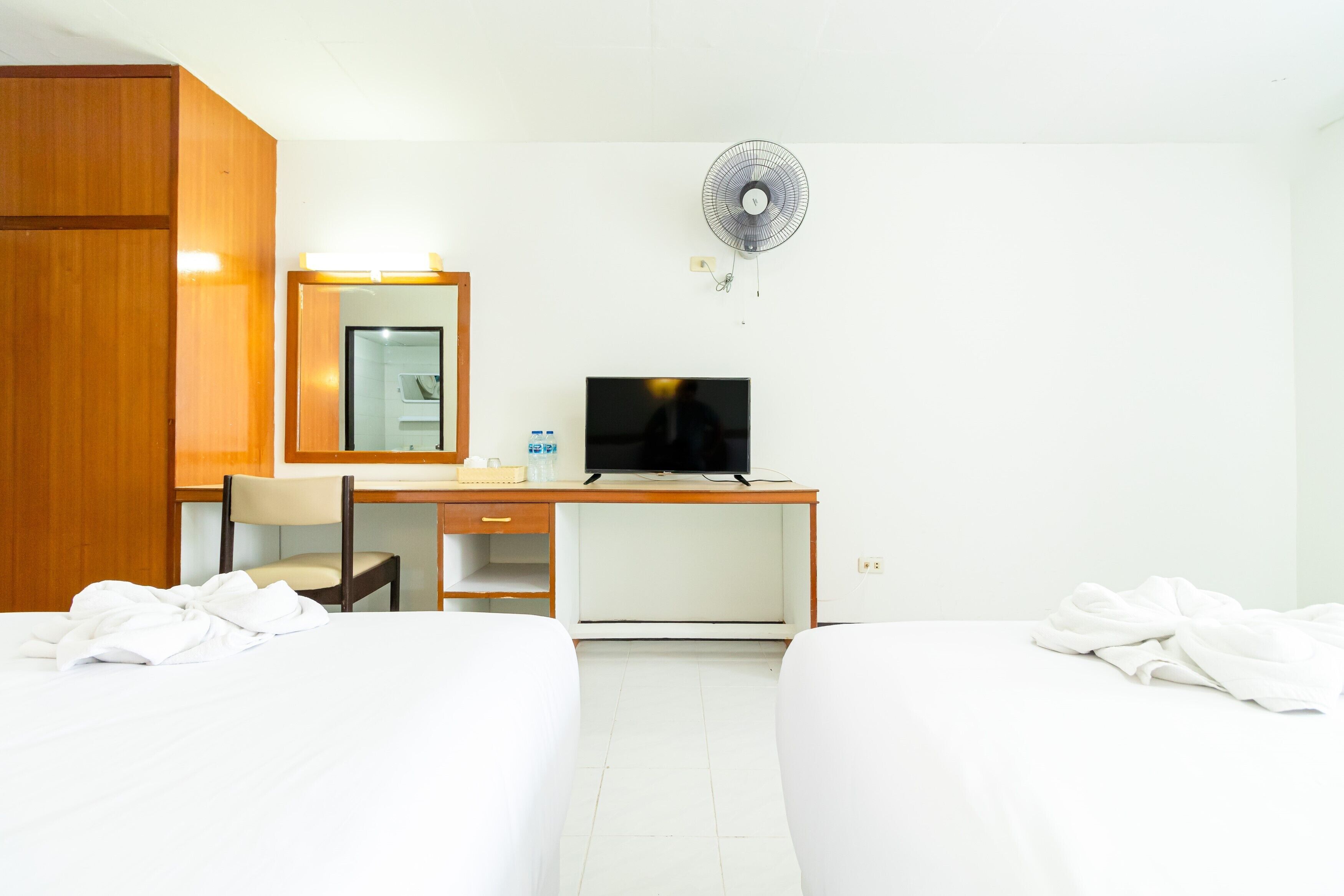 Habitación de hotel con dos camas, un escritorio con televisor, una silla, un espejo y un ventilador en la pared.