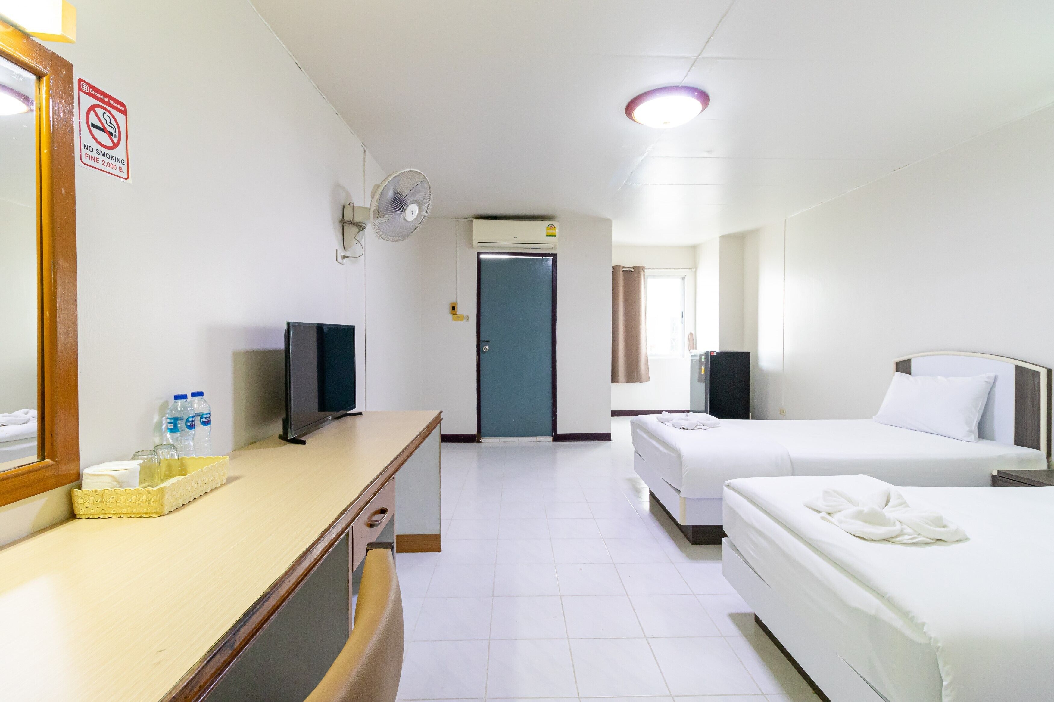 A hotel room with a bed, a desk with a mirror, a television, a fan, and an air conditioning unit.