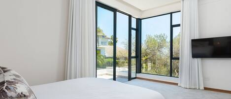 A modern bedroom with a large bed, a flat-screen TV, and a sliding glass door leading to a balcony with a view of the outdoors.