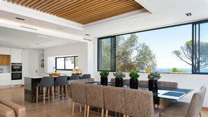 A modern kitchen with white cabinets, a dining area with a glass table and brown upholstered chairs, and a large window with a view of trees and the sea. - Coeur De Lion 5 (Cape Town)