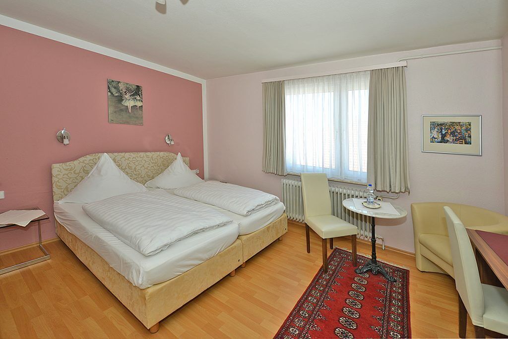 A bedroom with a double bed, a small round table with a chair, a radiator, and a window with curtains.