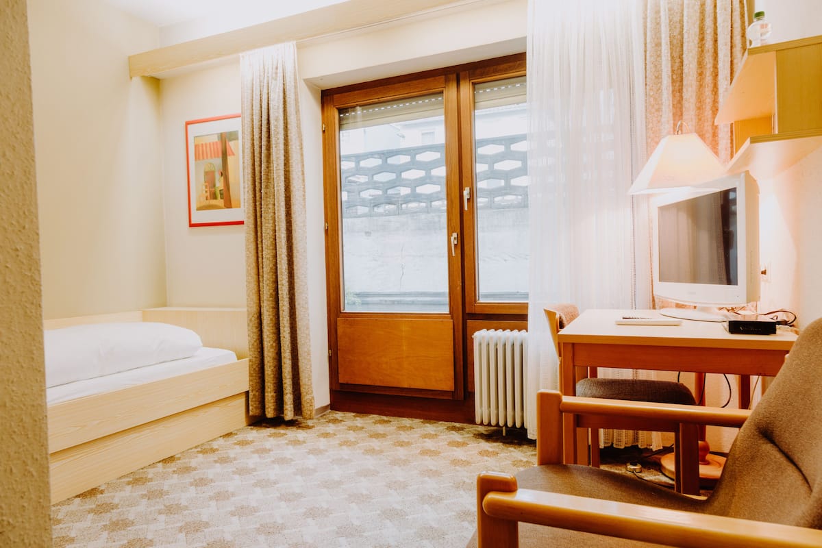 A hotel room with a bed, a desk with a computer, a chair, and a window with curtains.