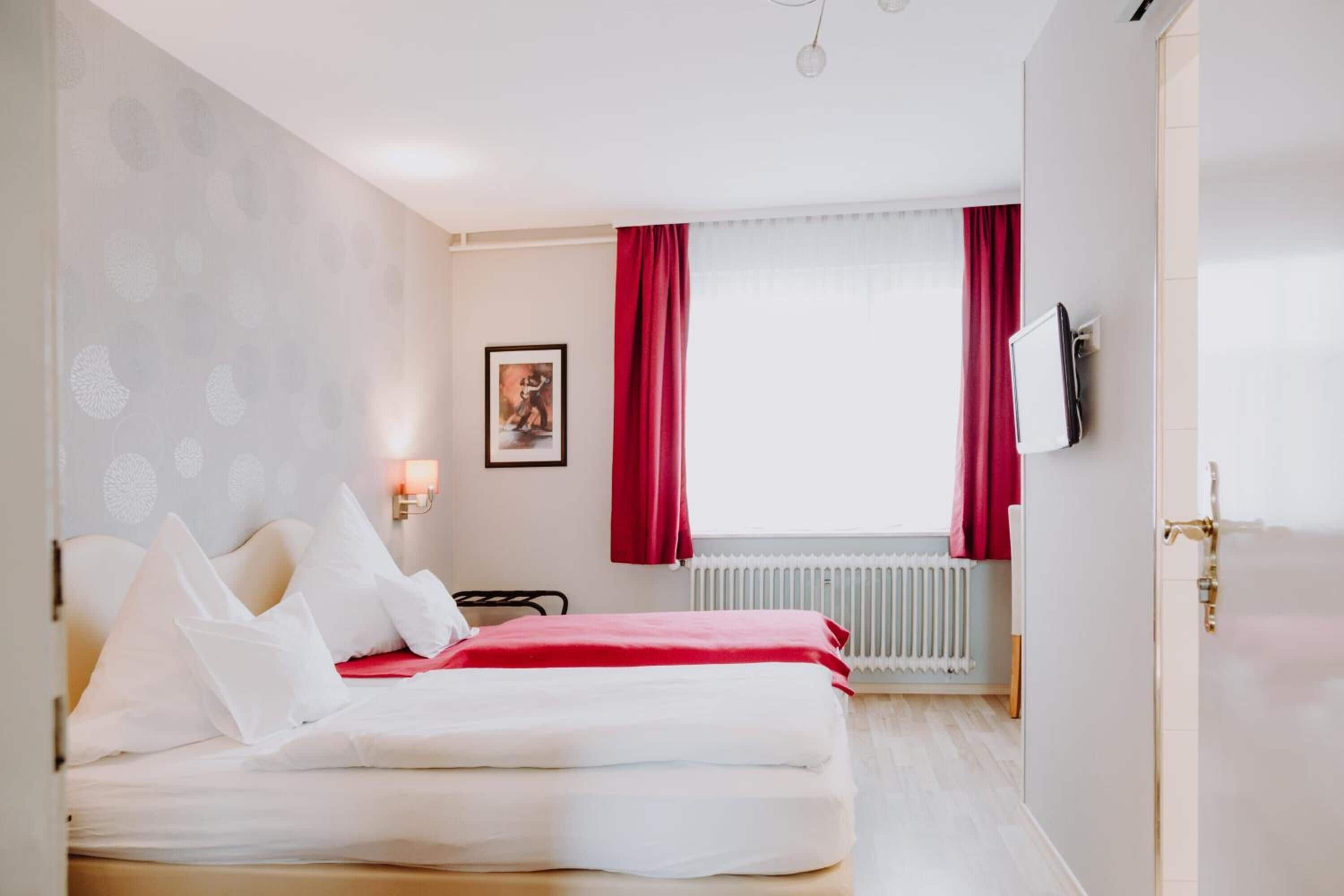 Habitación de hotel con cama, radiador, televisor montado en la pared y ventana con cortinas rojas.