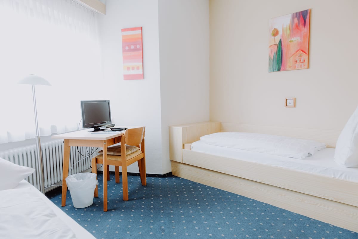 A hotel room with a bed, a desk with a computer, a chair, a radiator, and a wall with a painting.