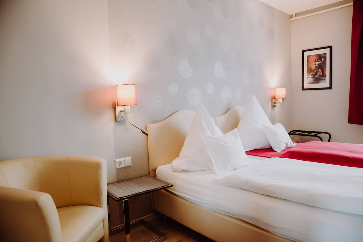 A hotel room with a bed, a chair, a nightstand, and wall-mounted lamps.