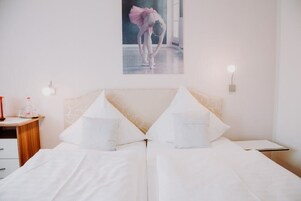 Quarto de hotel com duas camas, uma mesa de cabeceira de madeira, uma mesinha e uma luminária na parede.