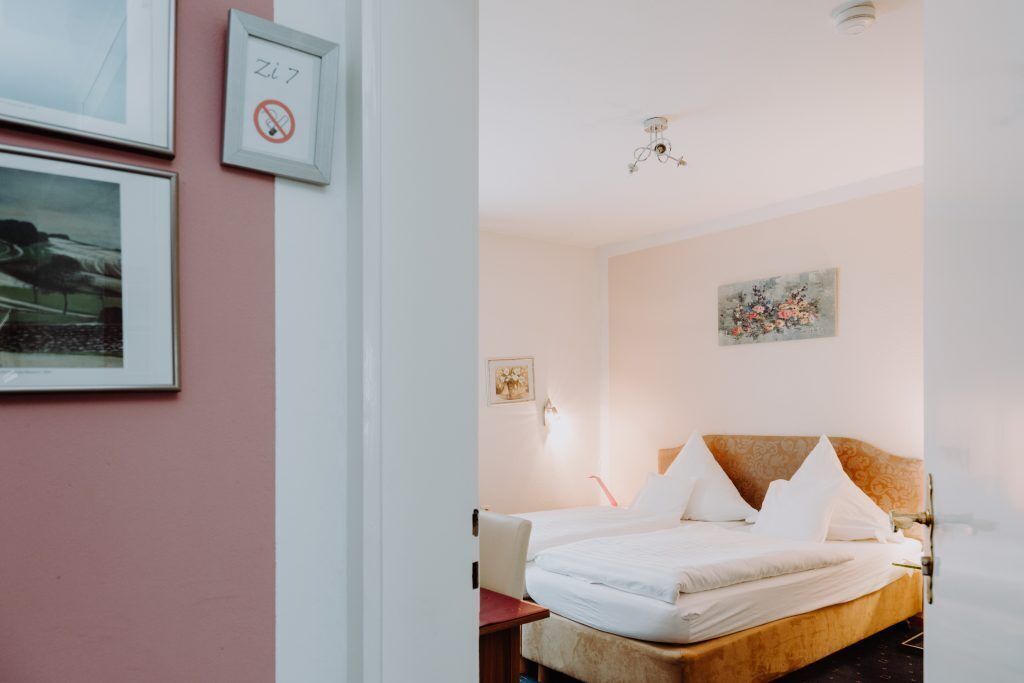 Quarto de hotel com uma cama, uma escrivaninha e uma cadeira. Há quadros na parede.