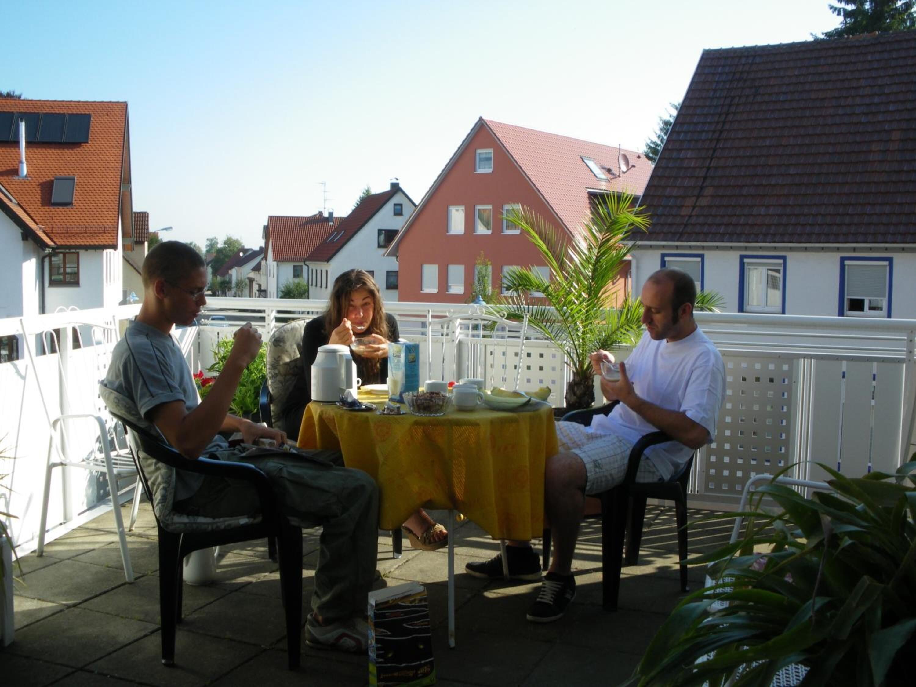 Three people seated at an outdoor table with a yellow tablecloth, surrounded by a white fence and houses in the background.