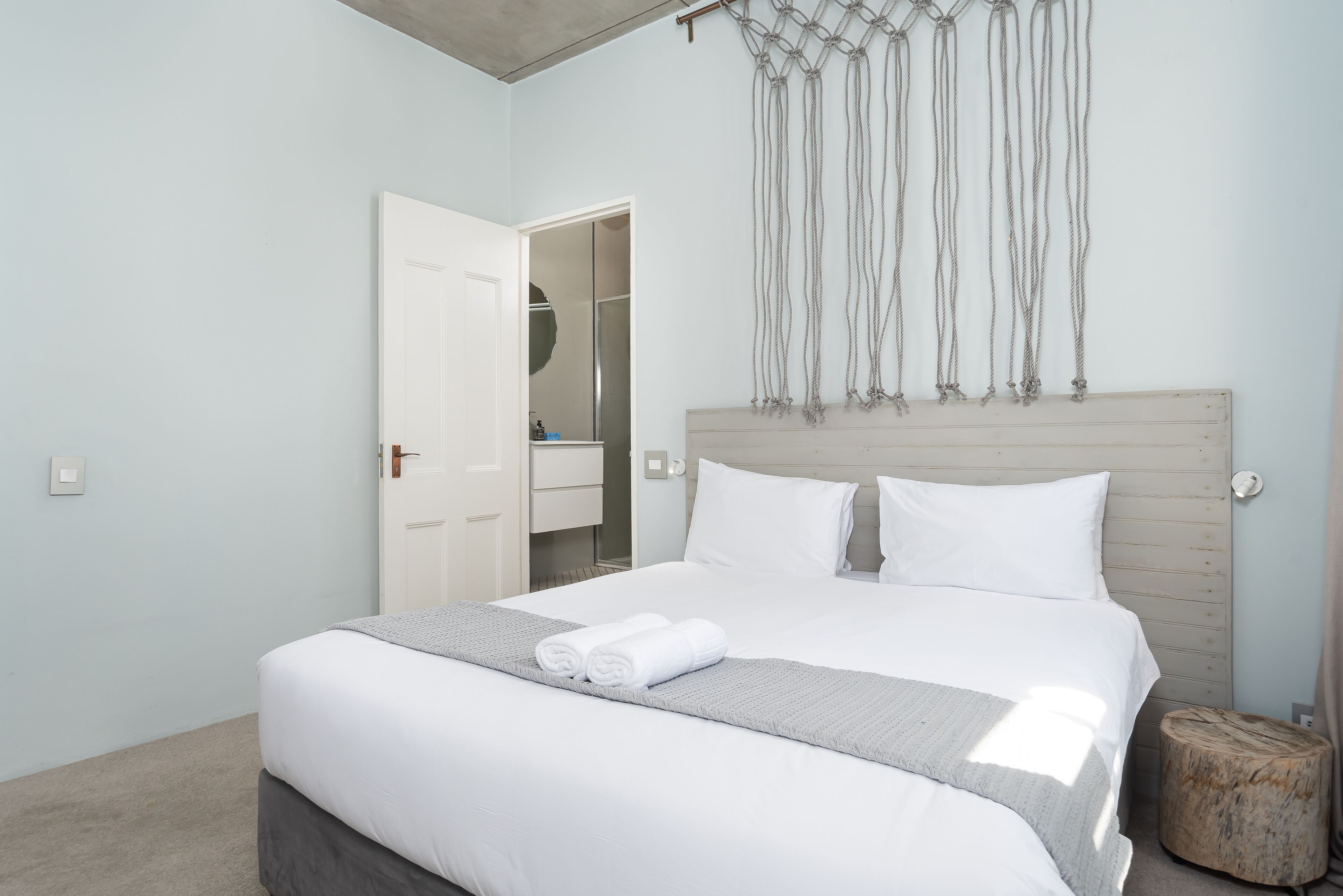 Ein ordentlich bezogenes Bett mit weißen Laken und einer grauen Decke, ein Holz-Kopfteil und ein Wandbehang aus Fransen.