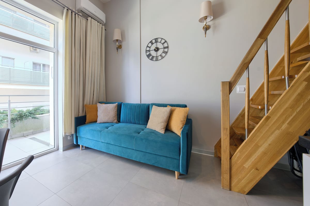 Una sala de estar moderna con un sofá azul, una mesa de comedor con sillas, una escalera de madera y vistas al exterior.