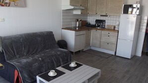 Un espacio habitable pequeño con un sofá gris, una mesita de centro blanca con dos tazas, una zona de cocina con microondas y refrigerador.