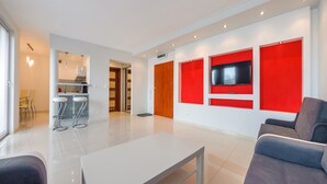 A modern living room with a white coffee table, grey sofa, and red accent walls. There is a TV mounted on the wall and a kitchen area in the background.