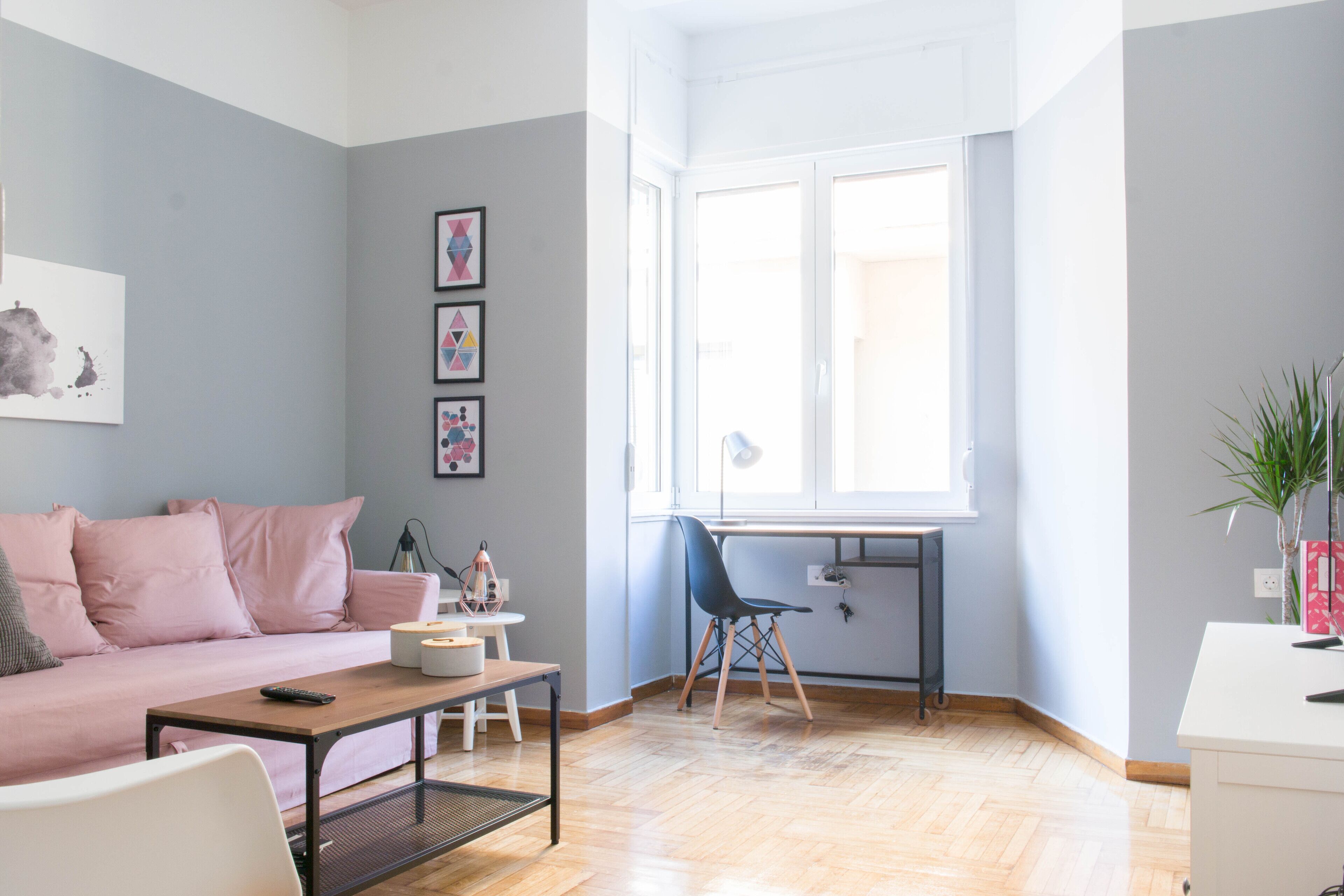 Een moderne woonkamer met een bank, salontafel en een bureau met stoel. Er hangen afbeeldingen in lijsten aan de muur en een raam zorgt voor natuurlijk licht.