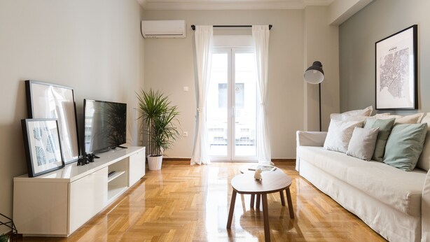 Un salon moderne avec un canapé blanc, une table basse, un téléviseur posé sur un meuble blanc, une plante en pot et une grande fenêtre avec des rideaux.