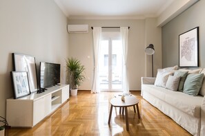 A modern living room with a white sofa, a coffee table, a TV on a white stand, a potted plant, and a large window with curtains. - UPSTREET Sleek Flat in Central Syntagma (Athens)