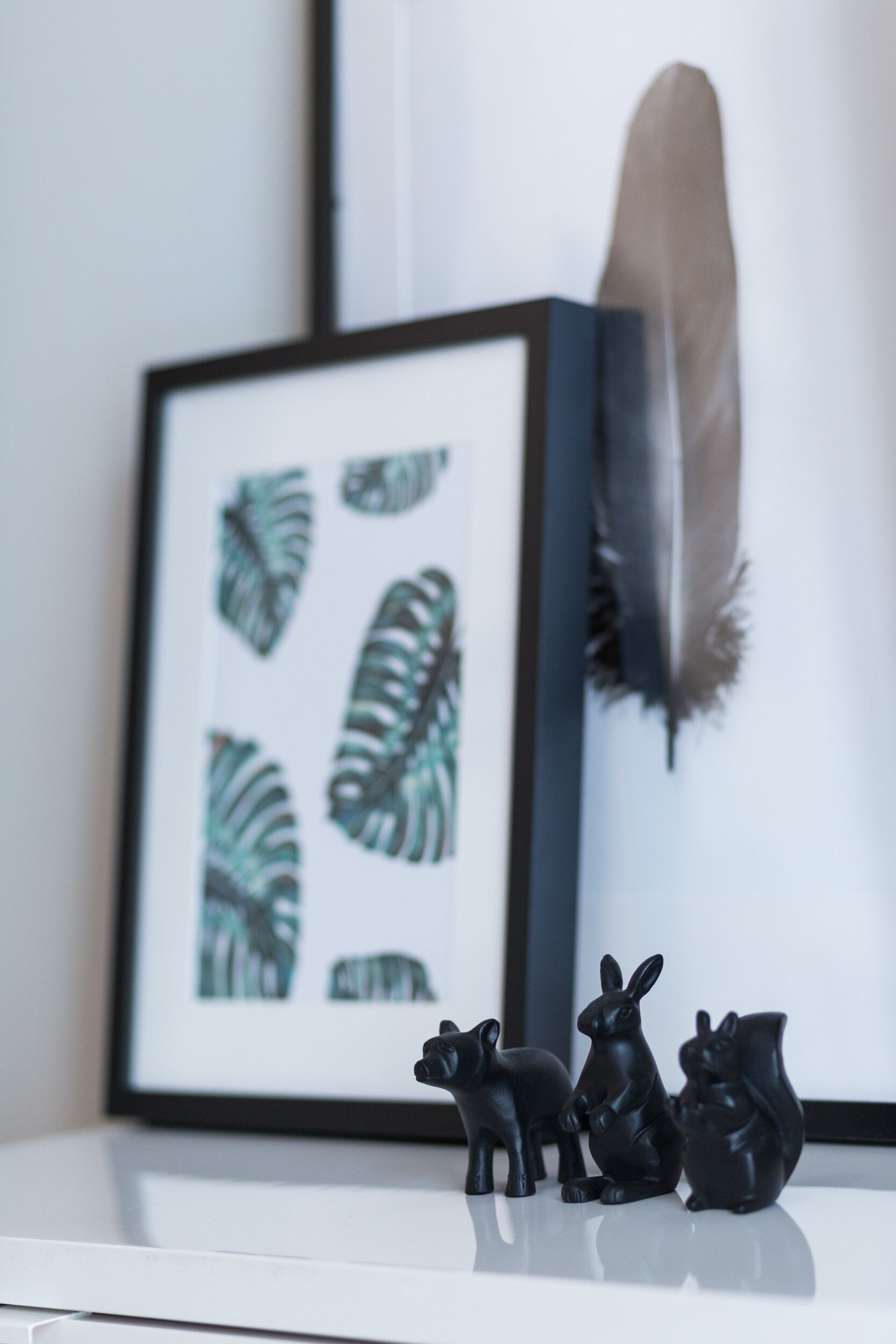 A framed artwork with tropical leaf patterns, a feather, and three black animal figurines on a white surface.