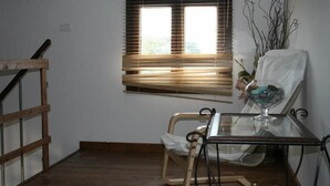 A room with a wooden floor, a window with blinds, a chair, a small table with a glass top, and a decorative vase. - Alba e tramonto B&B (Mola di Bari)