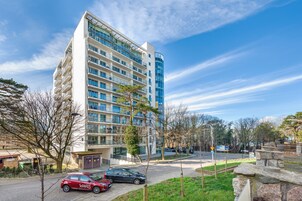 A modern multi-story building with balconies, a parking area, and a clear sky.