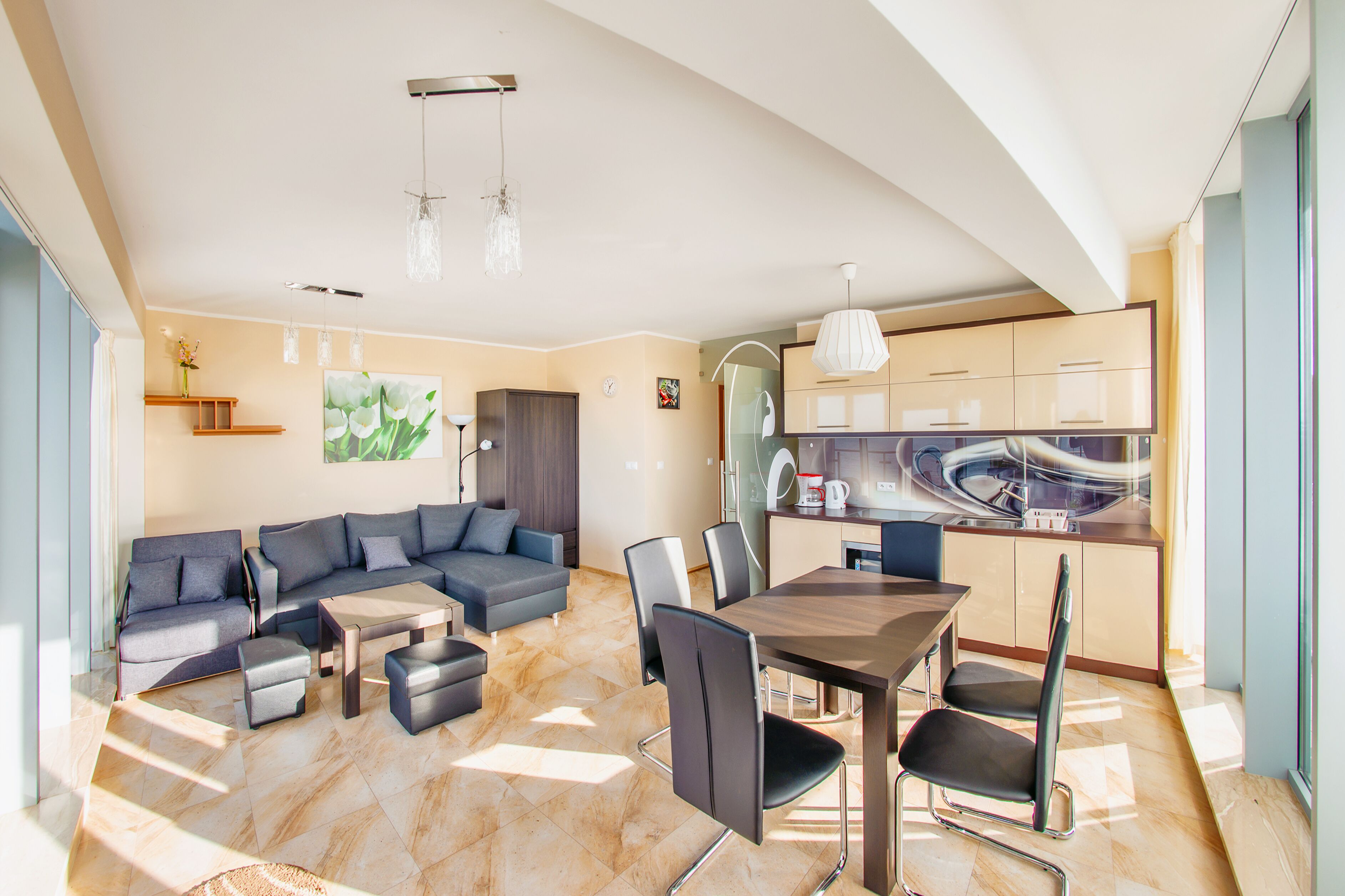 A modern living room with a sectional sofa, a dining table with chairs, and a kitchen area with cabinets and a coffee machine.