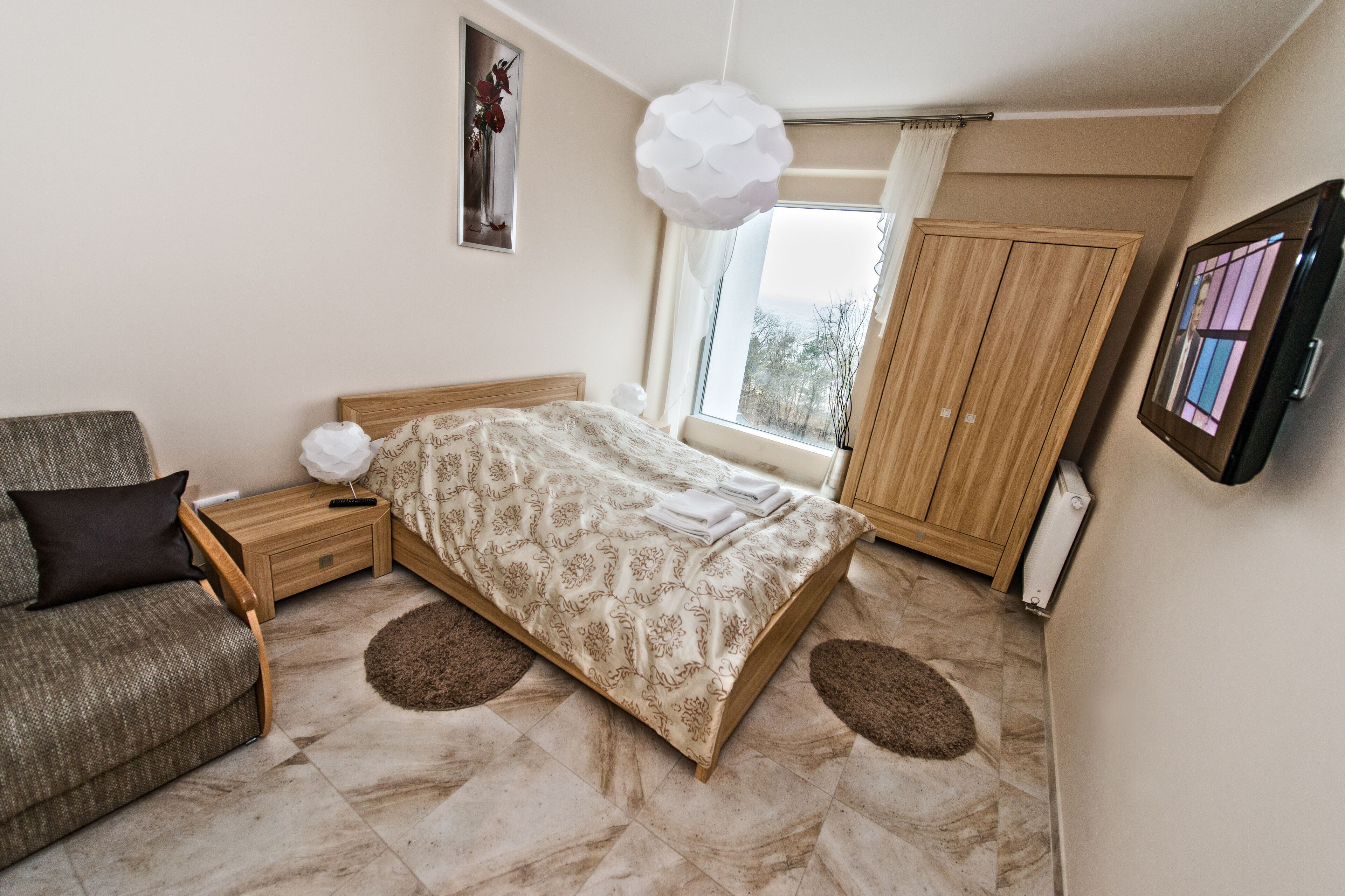 A bedroom with a bed, a wooden nightstand, a chair, a large window, and a wardrobe.