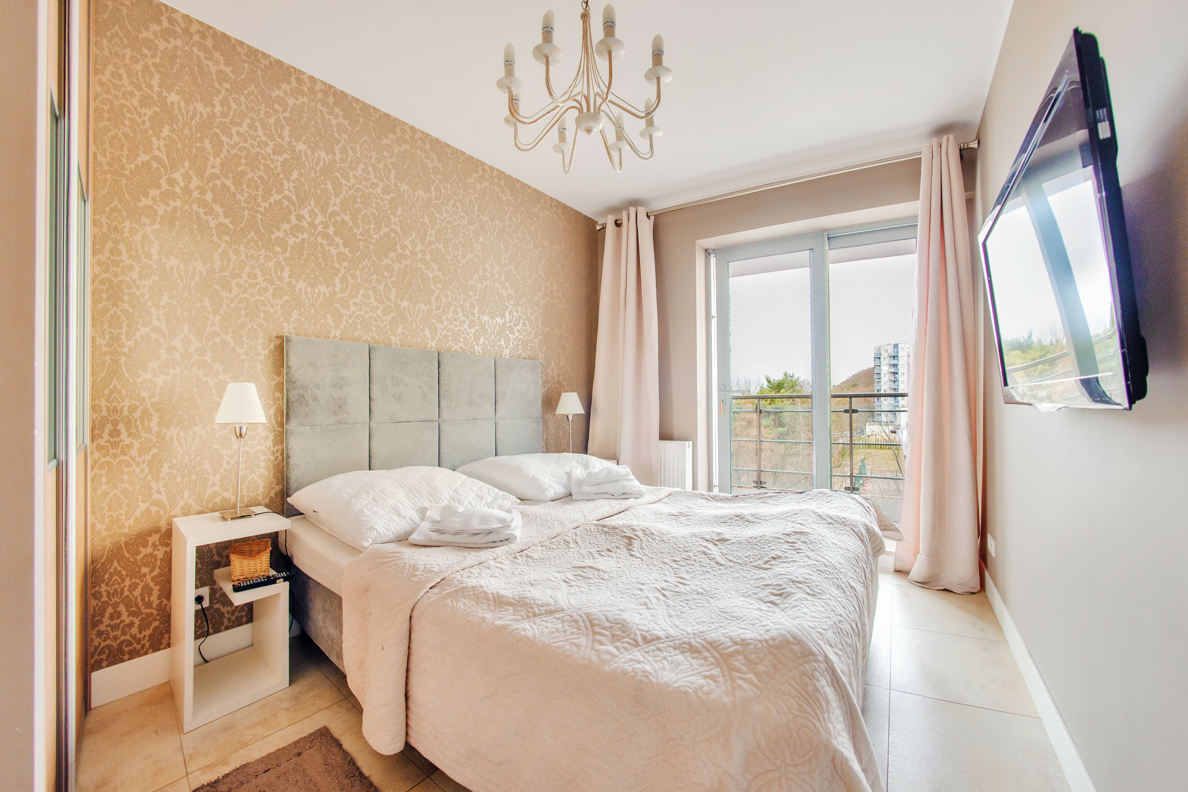 A bedroom with a large bed, a nightstand, a lamp, a TV mounted on the wall, and a view of the outdoors through a window.