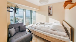 A modern bedroom with a bed, a chair, a large window, and a view of the city. - Sun & Snow Wechta - Promenada Gwiazd (Miedzyzdroje)