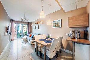Una sala moderna con un sofá, una mesa de comedor y sillas. Hay una zona de cocina con cafetera y utensilios. Una ventana ofrece vistas al exterior.