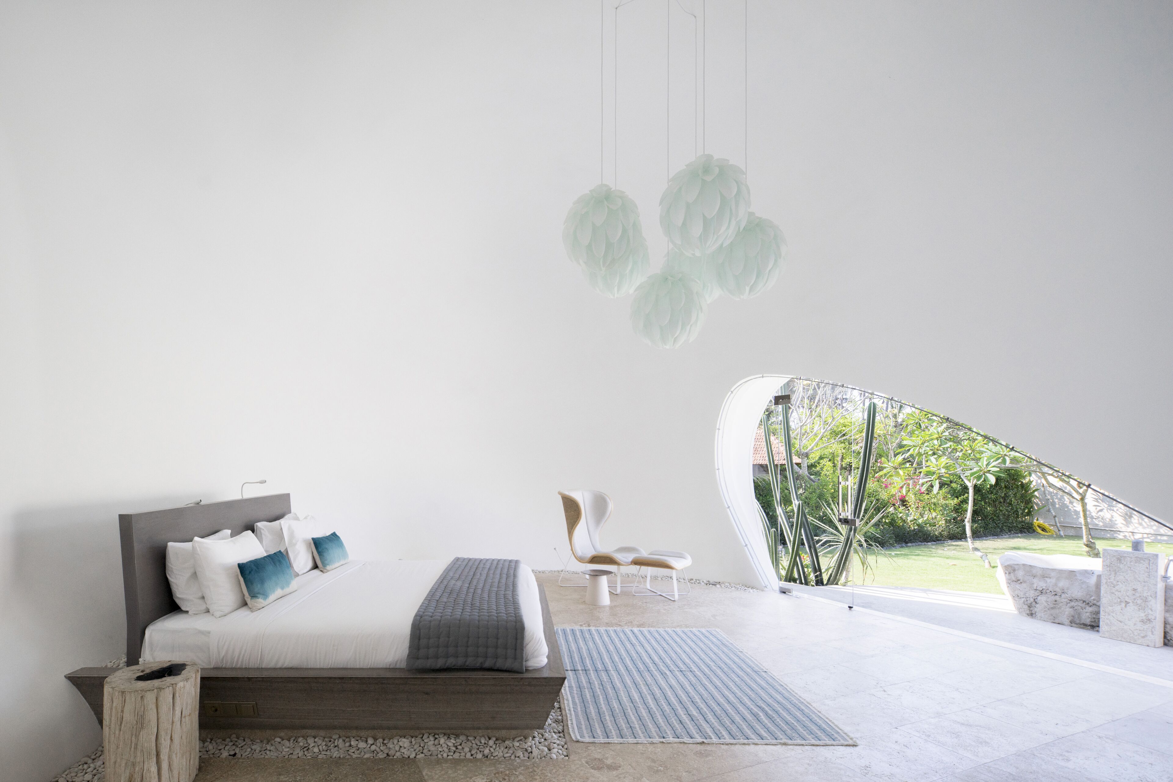 A modern bedroom with a bed, bedside table, and a chair. There is a view of a garden through a large window.