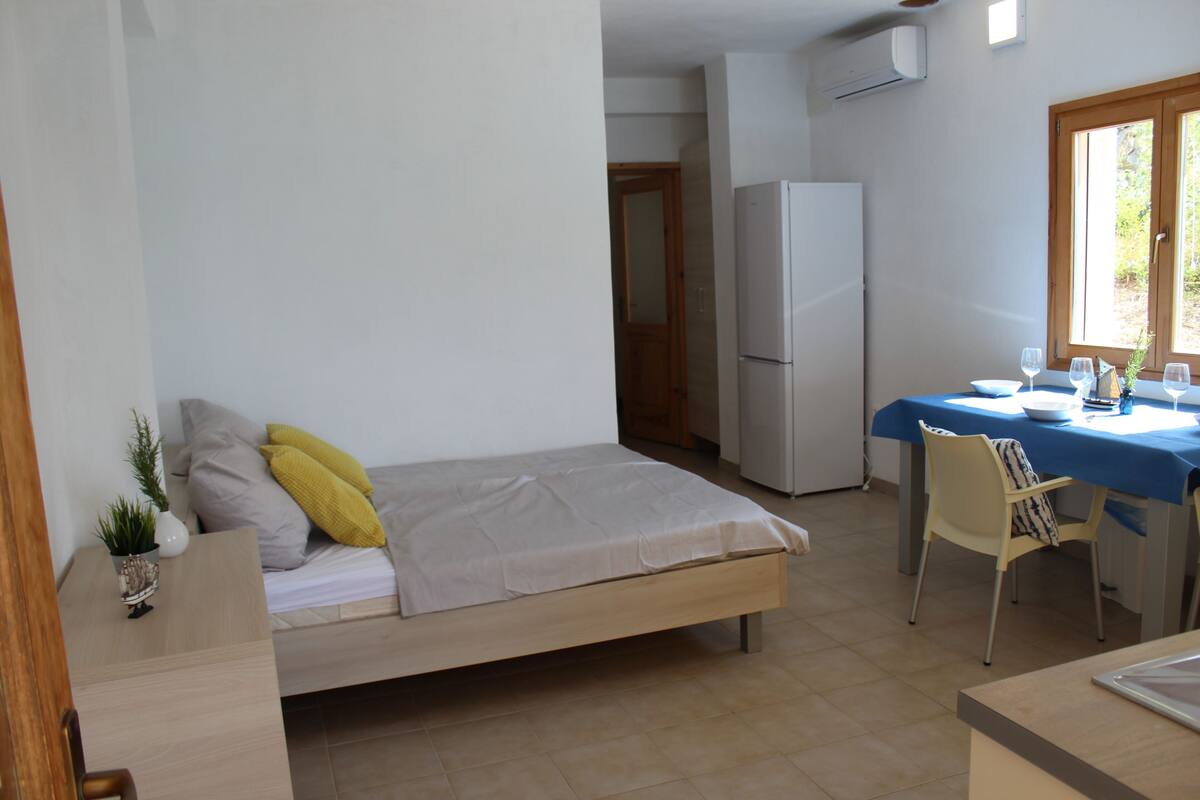 A compact room with a bed, a small table with chairs, a refrigerator, and a window.