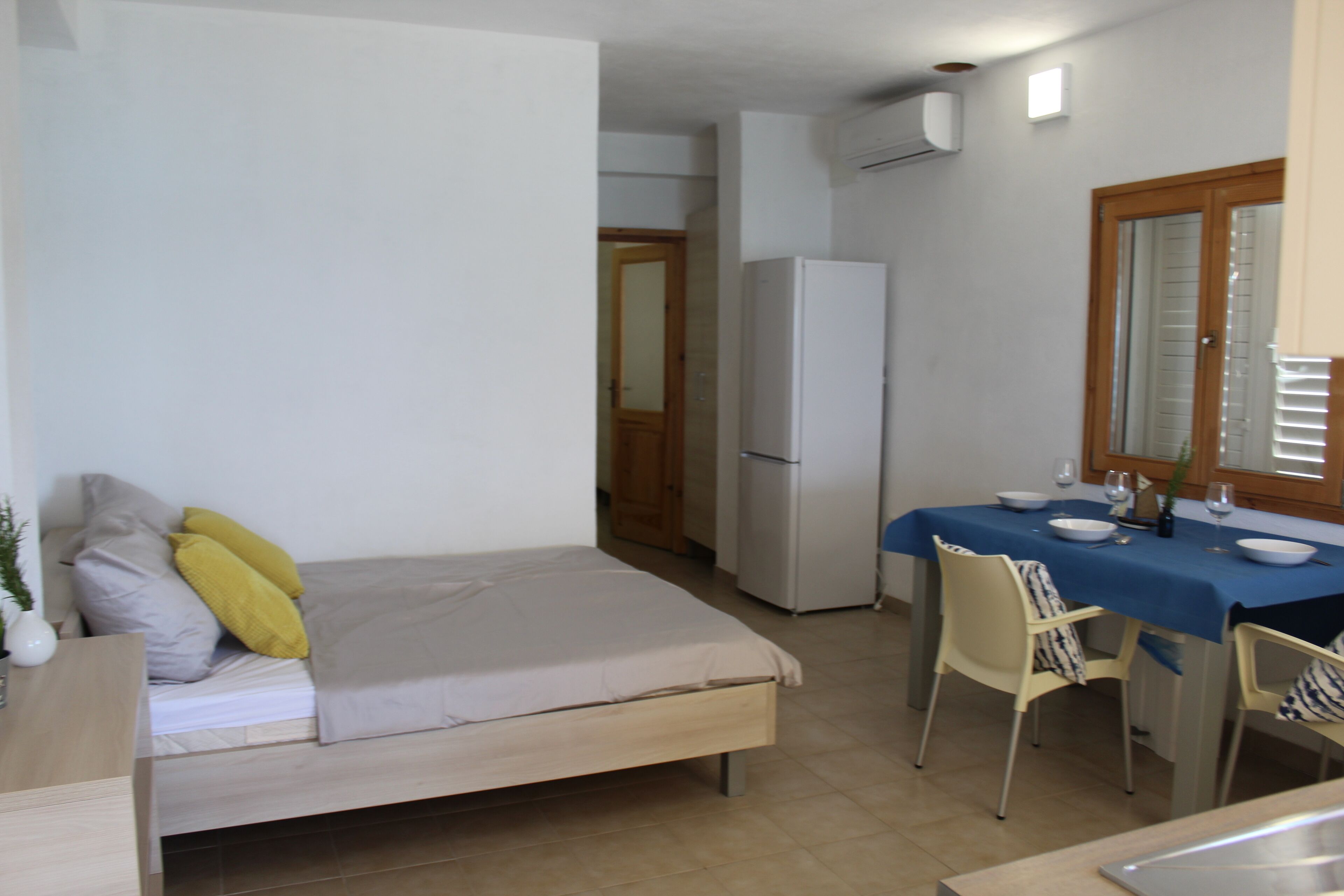 Ein kleines Zimmer mit einem Bett, einem Esstisch mit Stühlen, einem Kühlschrank und einem Fenster mit Jalousien.