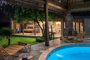 A poolside area with lounge chairs, a thatched roof, and a view of the interior. - Original Villa Pryaniki (Tabanan)