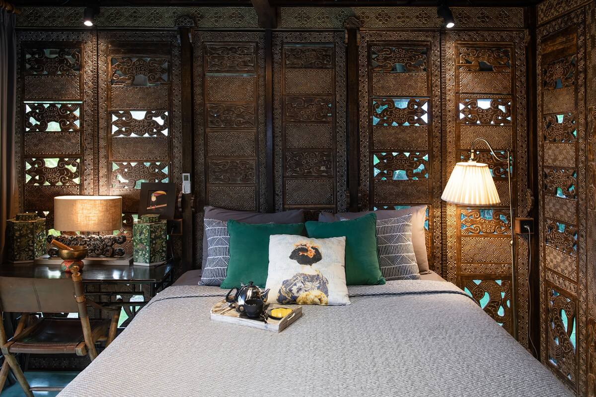 A room with a bed, pillows, a lamp, a table with a teapot, and a chair.
