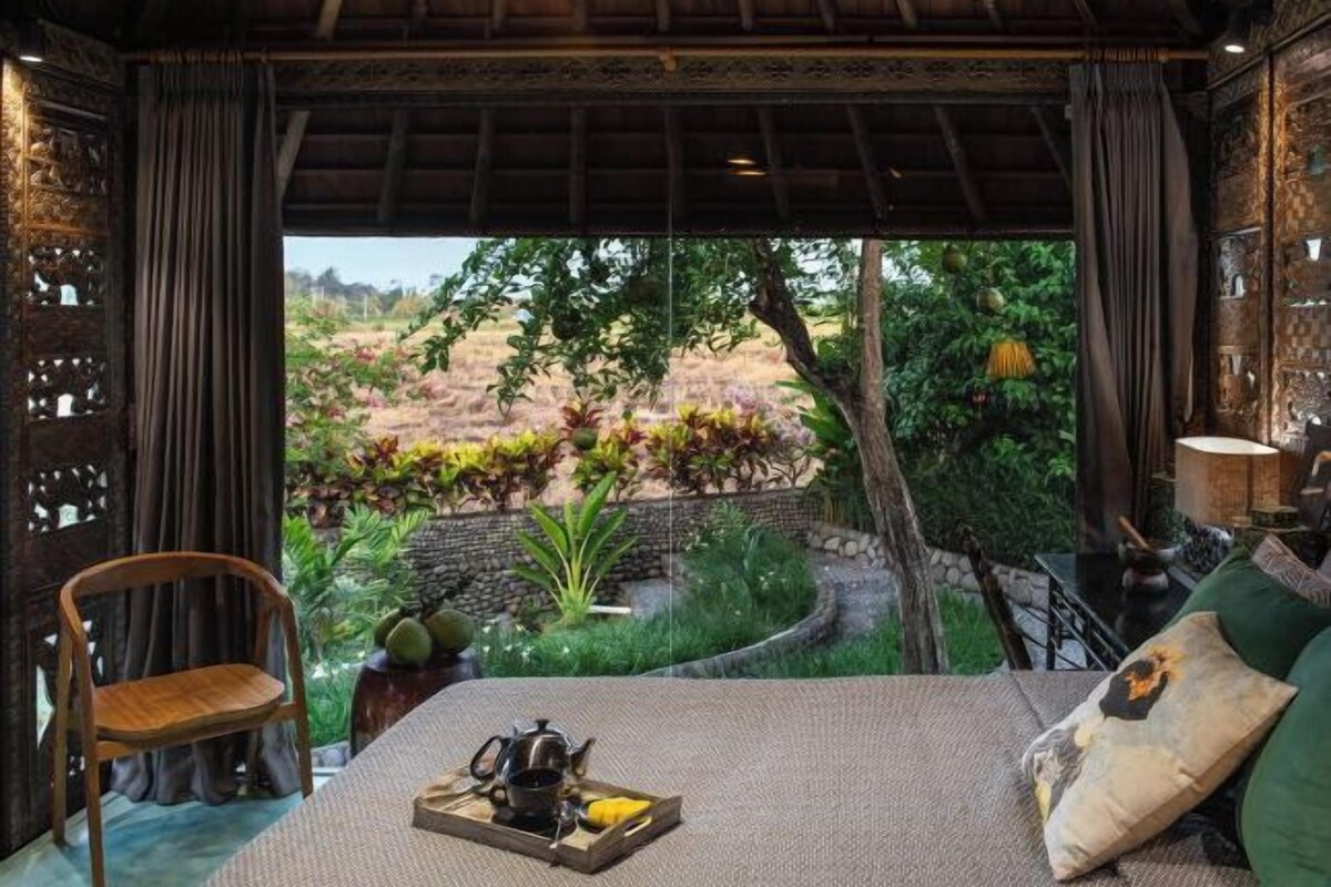 A room with a bed, a chair, a table with a tray of items, and a view of a garden.