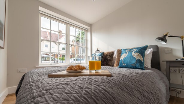 Een keurig opgemaakt bed met een grijze deken, een dienblad met croissants en jus d'orange, en een raam met uitzicht op huizen.