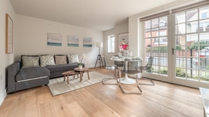 Een ruime woonkamer met een grijze bank, een klein rond tafeltje en een glazen eettafel met stoelen. Er zijn grote ramen met uitzicht op het omliggende landschap.