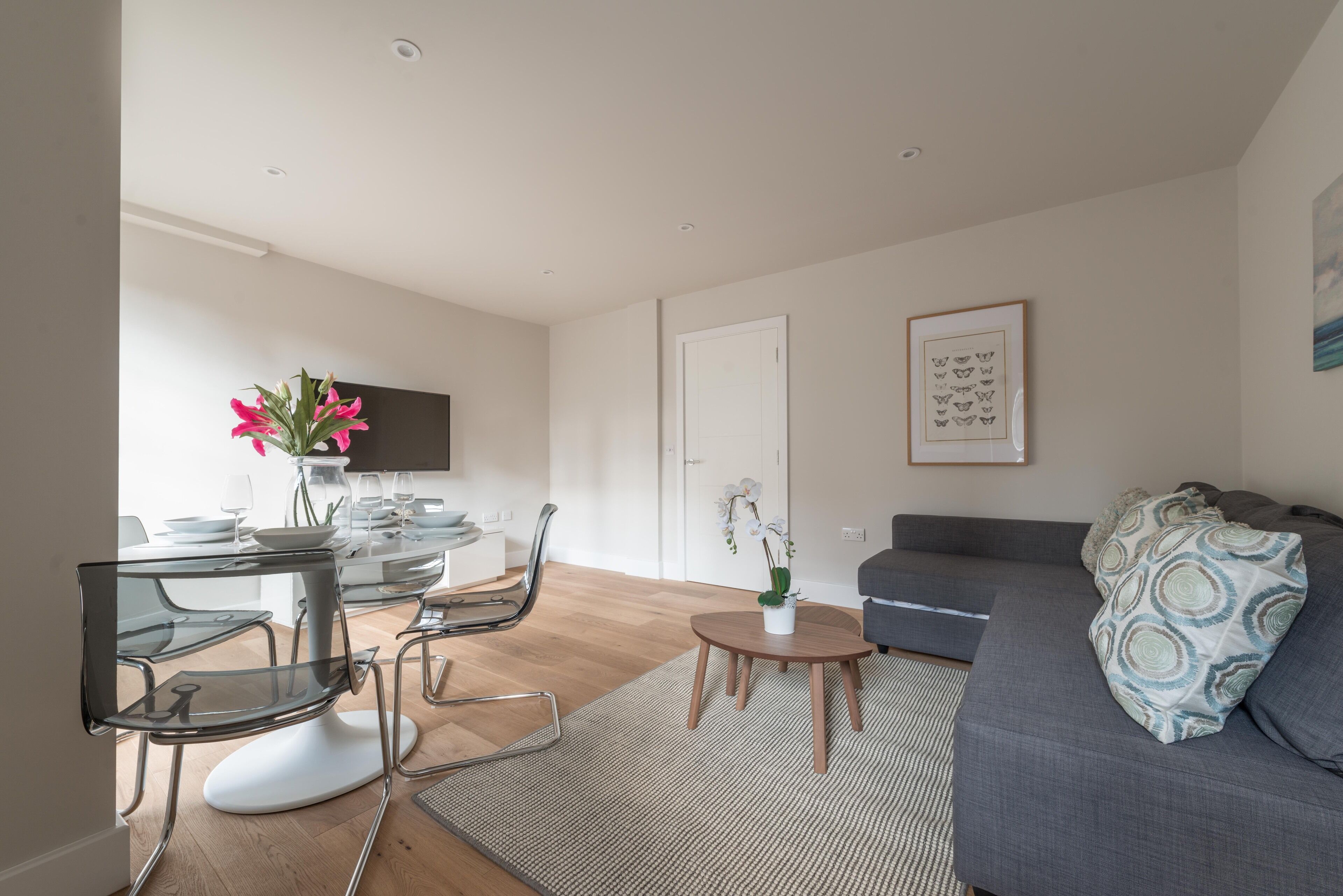 A modern living room with a grey sofa, a glass dining table with chairs, and a small round table with a vase of flowers.