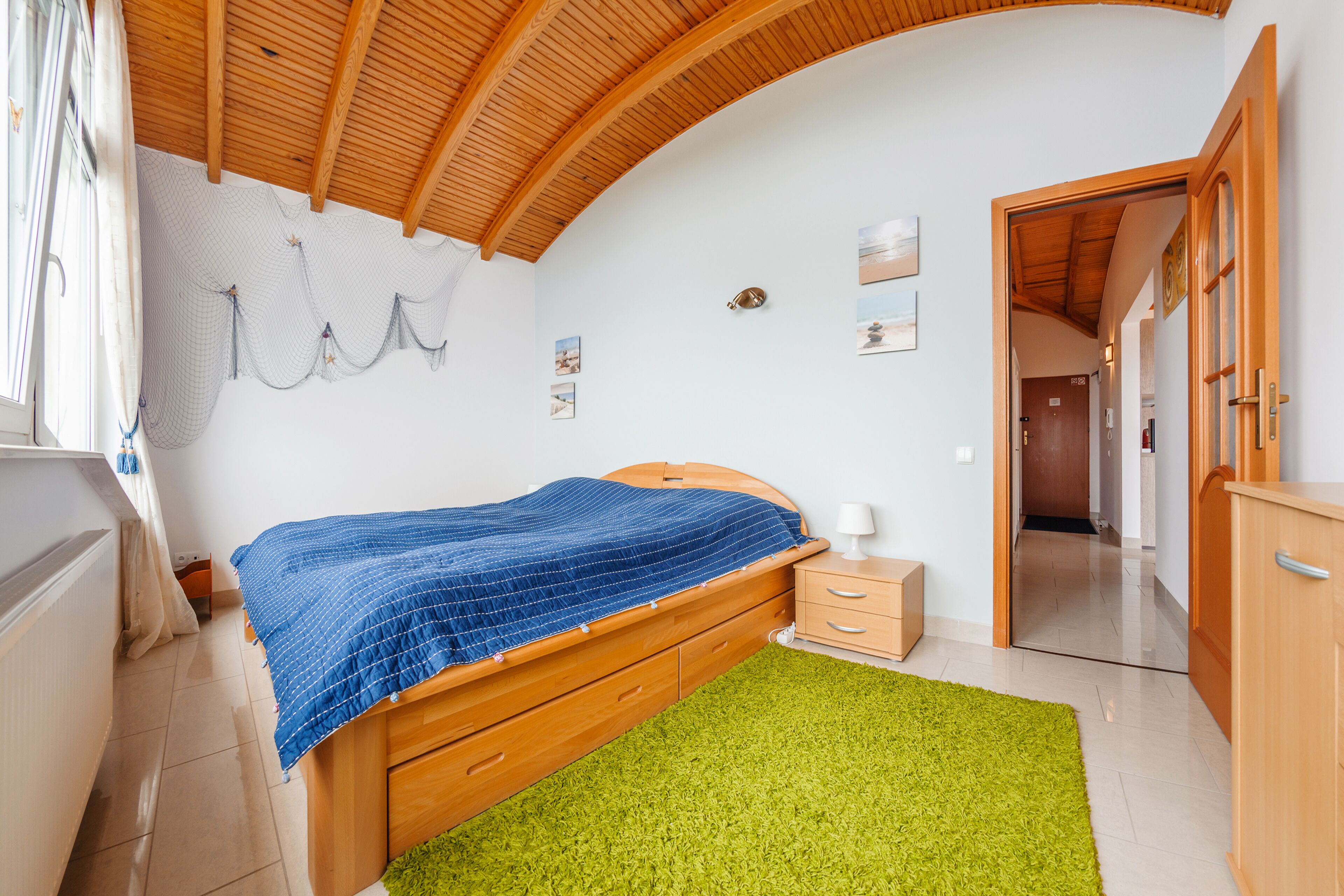 A bedroom with a wooden bed, a green rug, a wooden dresser, and a white wall with pictures.