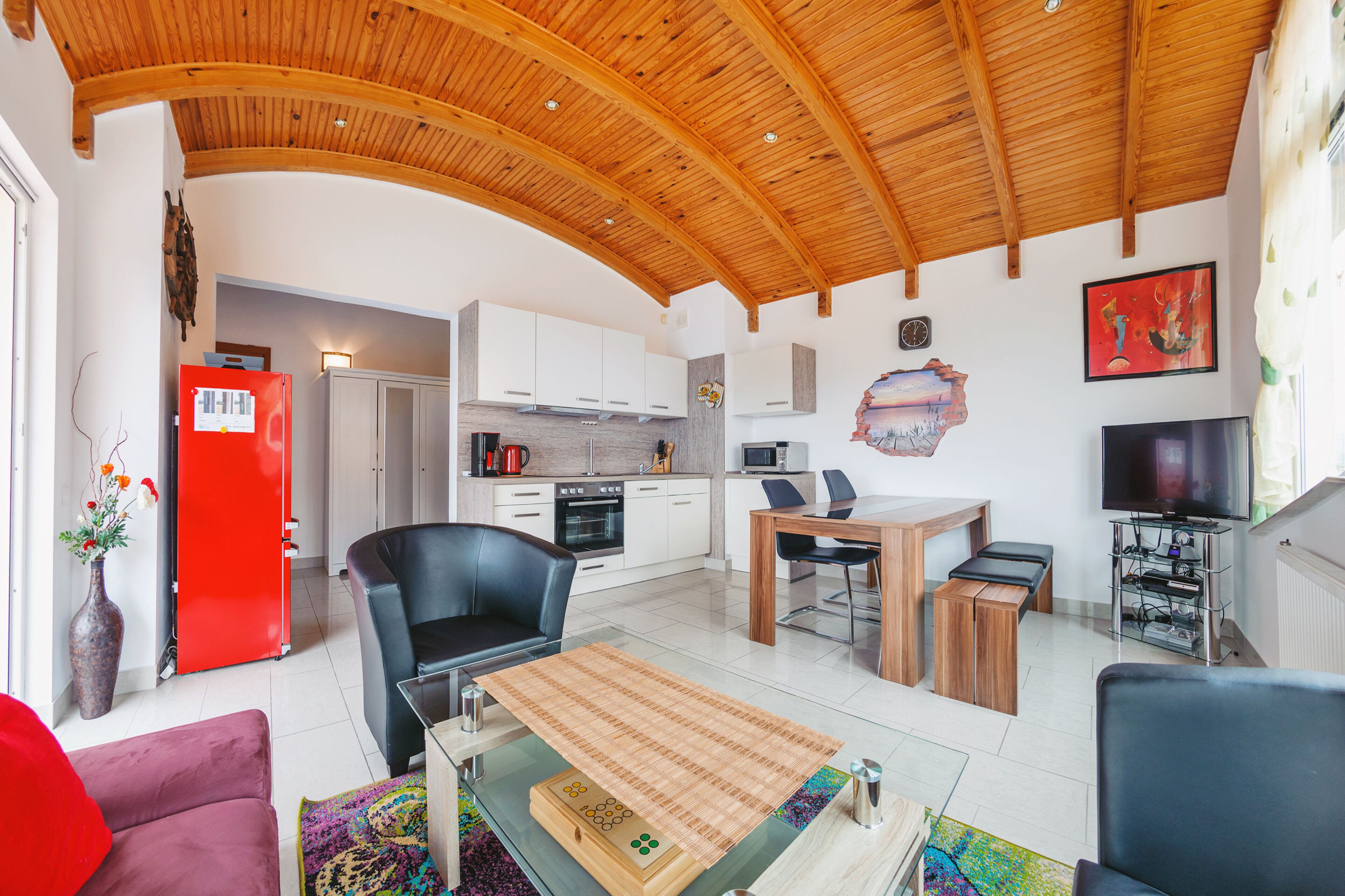 A spacious living area with a wooden ceiling, a kitchen with white cabinets, a dining table, and a TV.