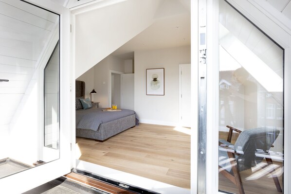 Un dormitorio moderno con techo inclinado, una cama con ropa de cama gris, una mesita de noche con lámpara y una obra enmarcada en la pared.