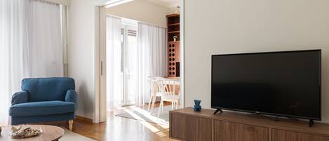 A living room with a blue armchair, a TV on a wooden cabinet, and a dining area with a table and chairs.
